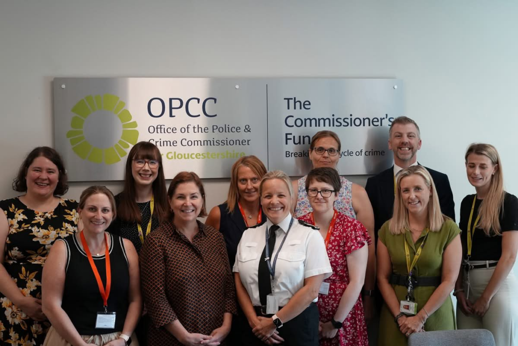 A group of people from the Gloucestershire Domestic Abuse Partnership and the Domestic Abuse Commissioner stand in front of a silver sign with blue and green designs.