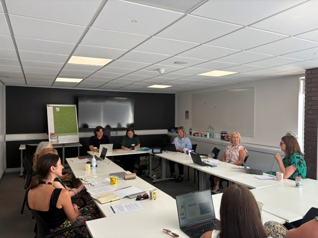 A group of people sit around tables arranged in a circle, with a flipchart and whiteboard one either side.
